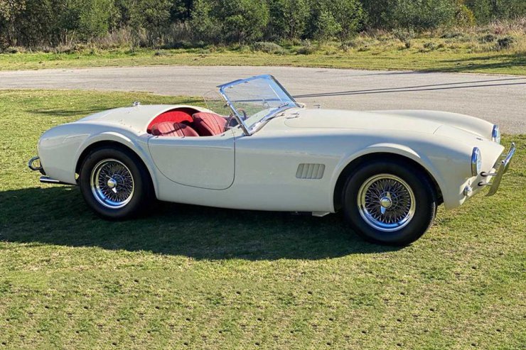 نمای جانبی رودستر ای سی شلبی کبرا / AC Shelby Cobra با صندلی قرمز و رنگ سفید در کنار جاده سرسبز